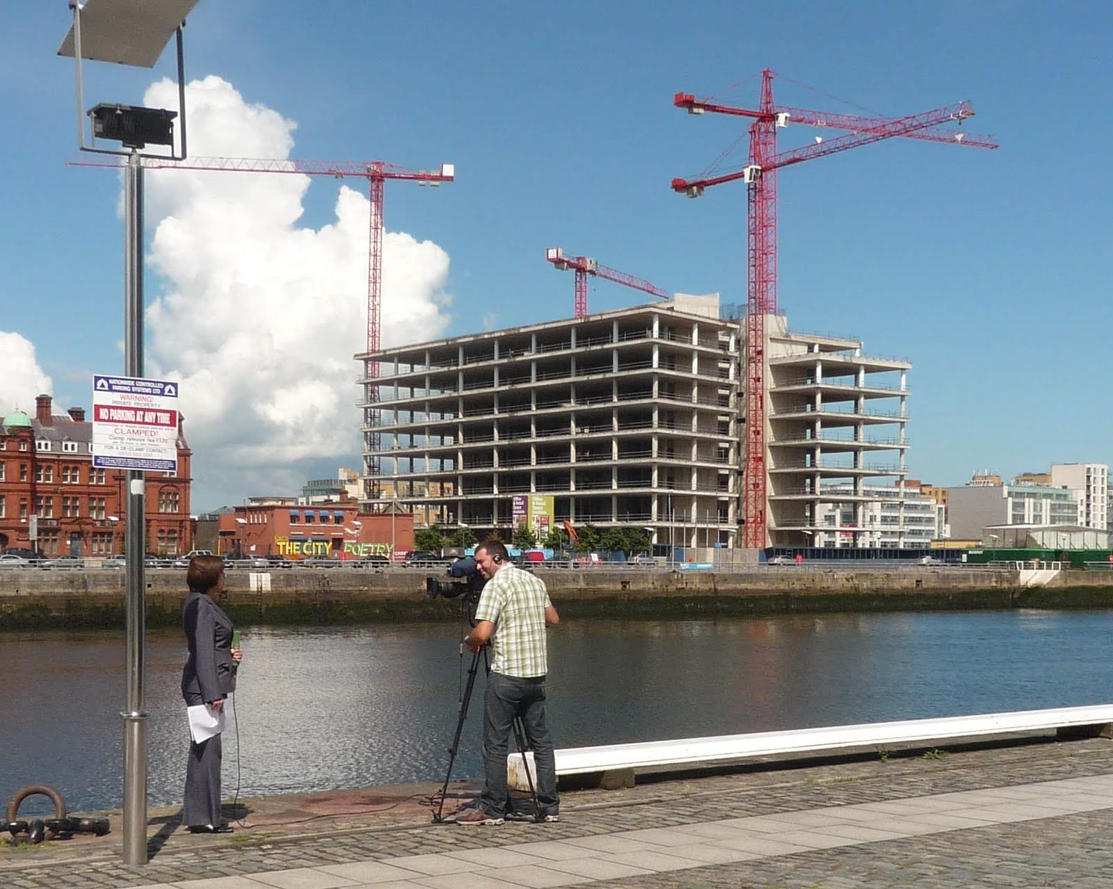 Colour photograph of the River Liffey showing the new Anglo Irish Bank Headquarters in the process of construction and surrounded by red cranes; there is a woman being filmed by a photographer in the foreground.