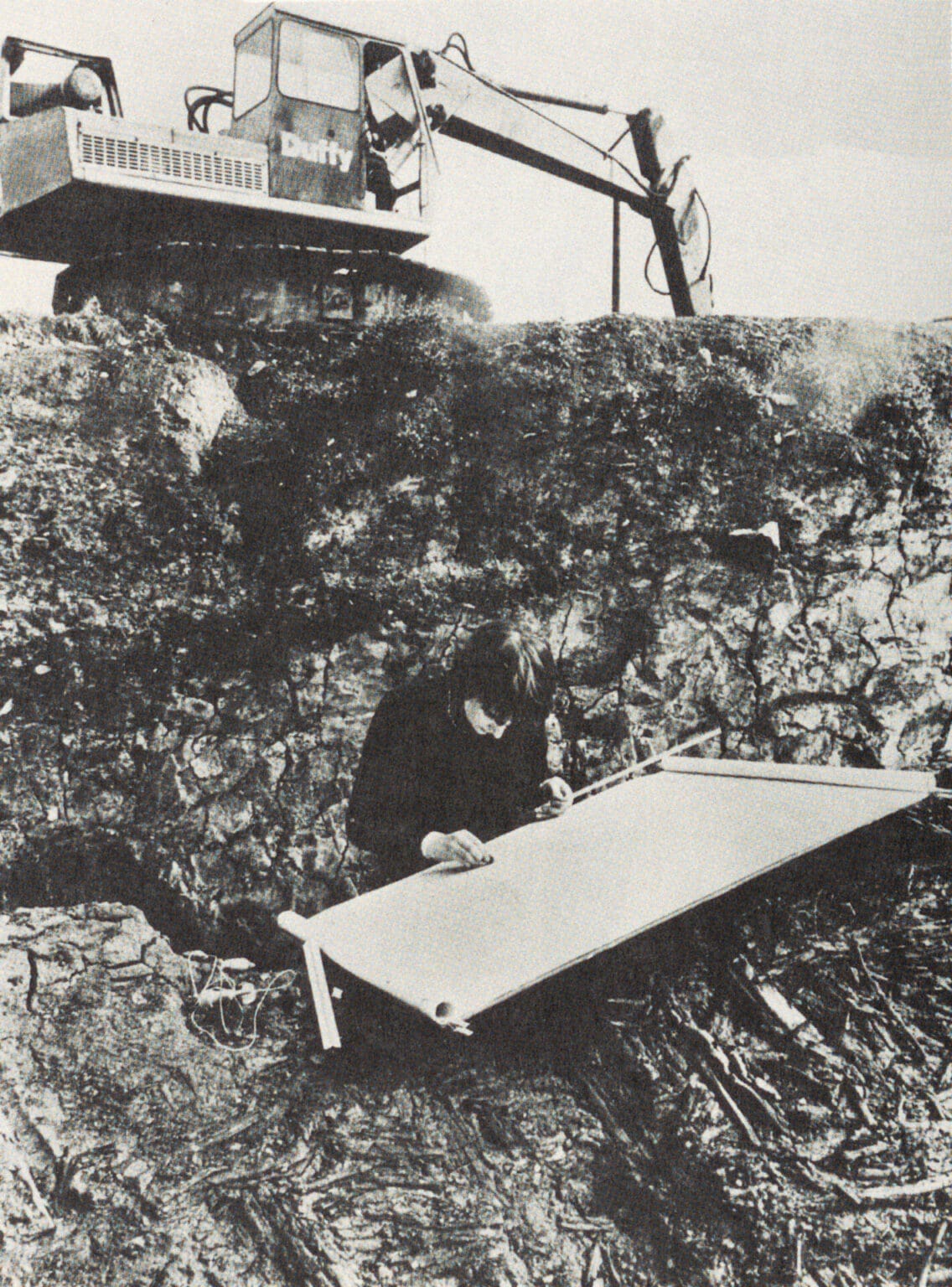 A black and white photograph taken in 1975 by Tom Lawlor and reproduced in the Irish Times showing architectural student Helen Lane records on paper the medieval structures of Wood Quay, Dublin. Behind her, a construction excavator is clearing the land.