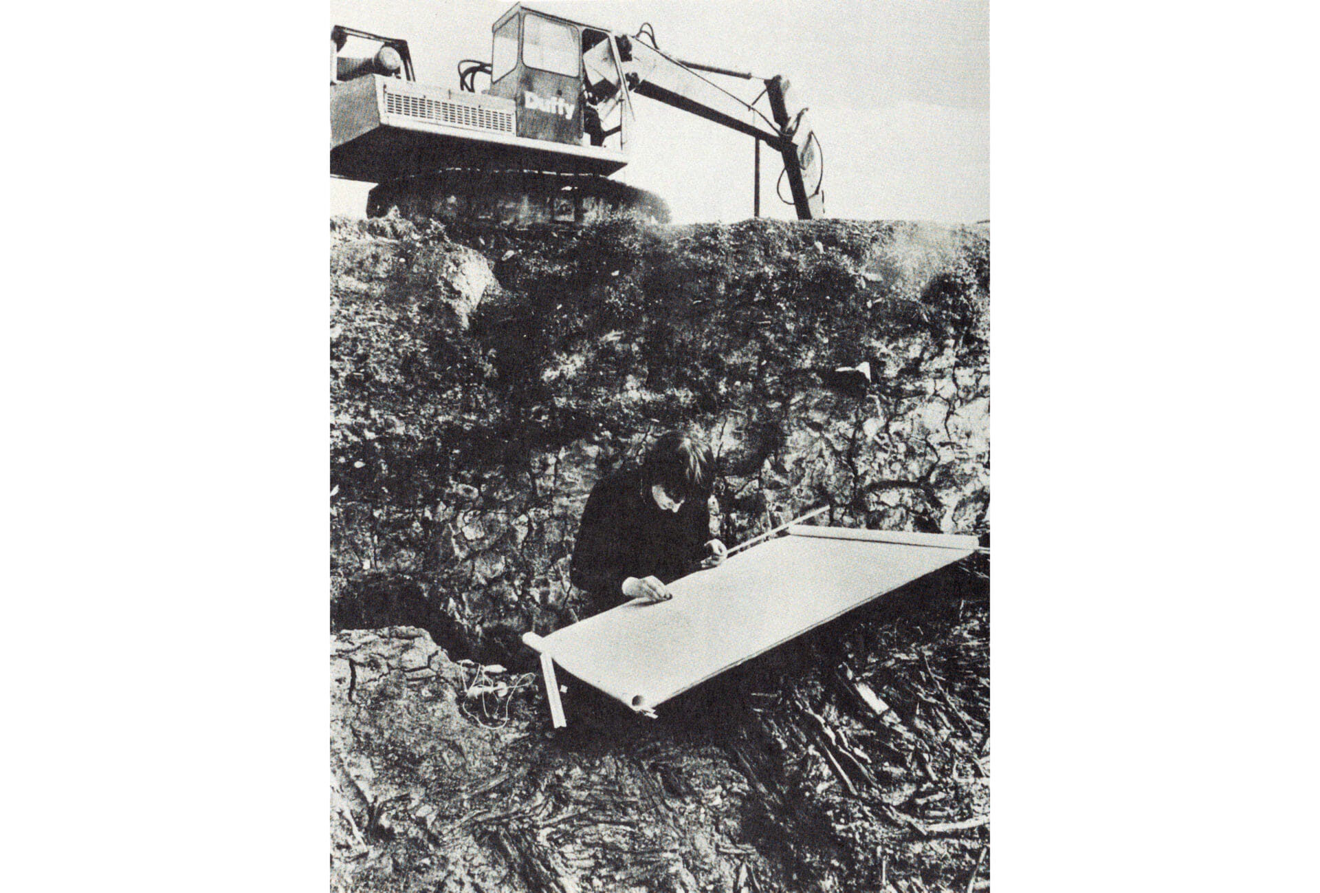 A black and white photograph taken in 1975 by Tom Lawlor and reproduced in the Irish Times showing architectural student Helen Lane records on paper the medieval structures of Wood Quay, Dublin. Behind her, a construction excavator is clearing the land.